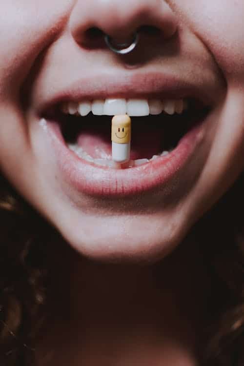 Close-up of a woman holding a pill with a happy face emoji between her teeth, symbolizing mood stabilizers for mental health.