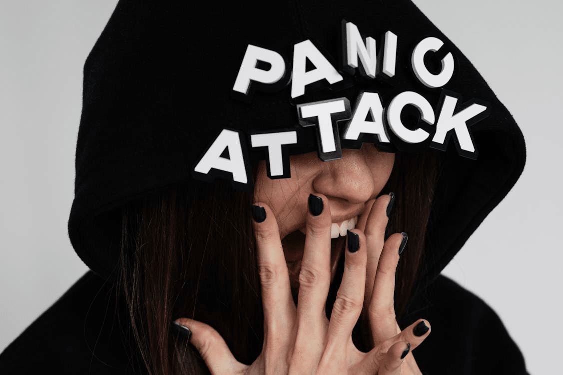 Woman wearing black hoodie with "panic attack" written on top, covering her face and appearing to scream.
